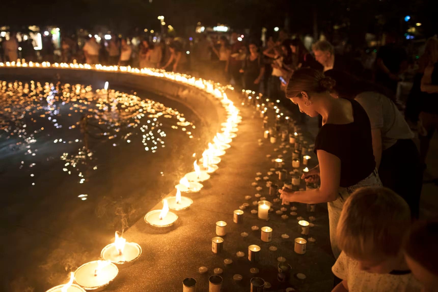 Mourners gathered at a nighttime vigil, one leaning over to light a candle among memorial signs